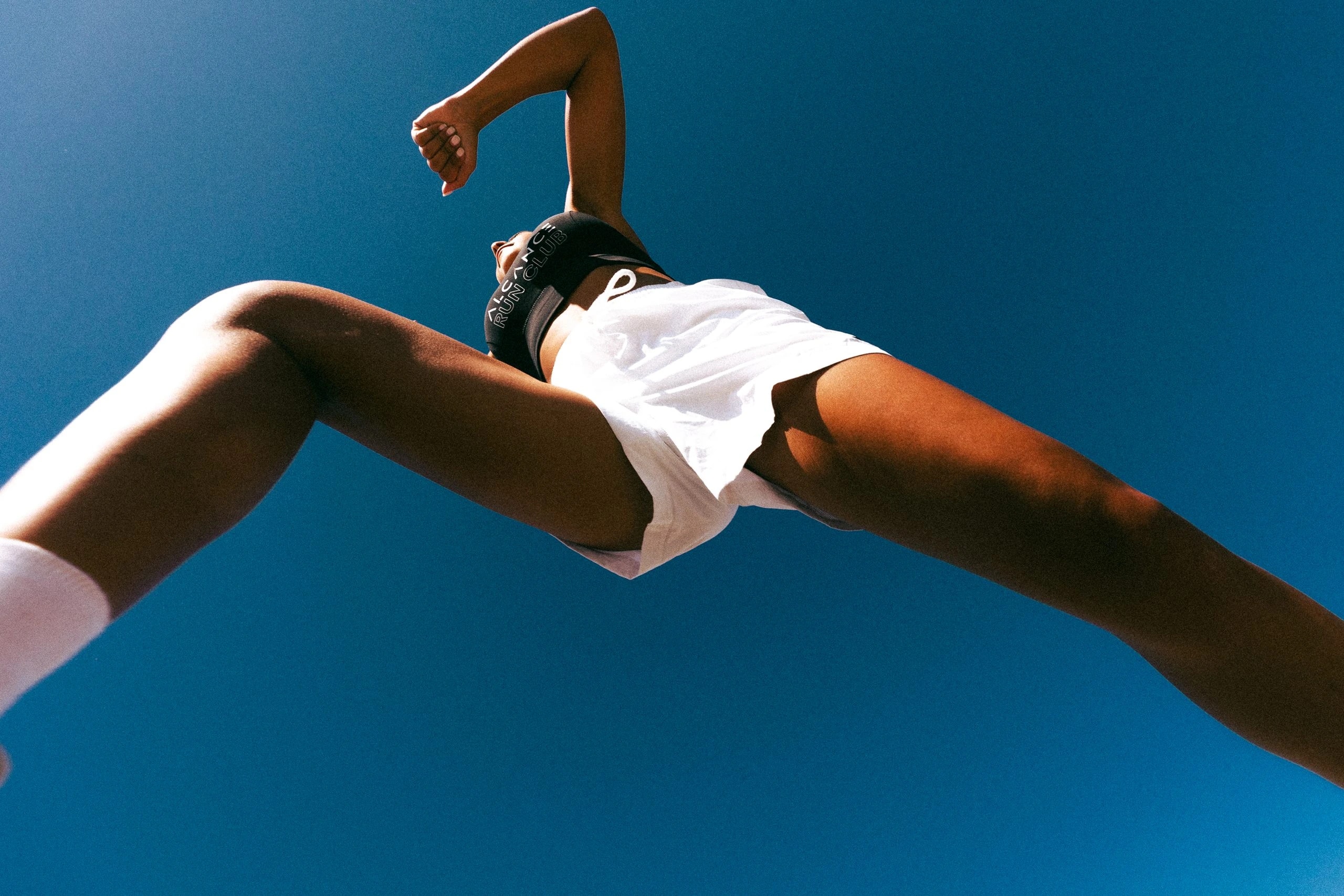 An Alcance Run Club runner mid-stride, shot from below against a clear blue sky